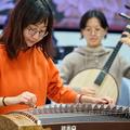 A woman in an orange jumper plays a guzheng; a woman in the background plays a stringed instrument