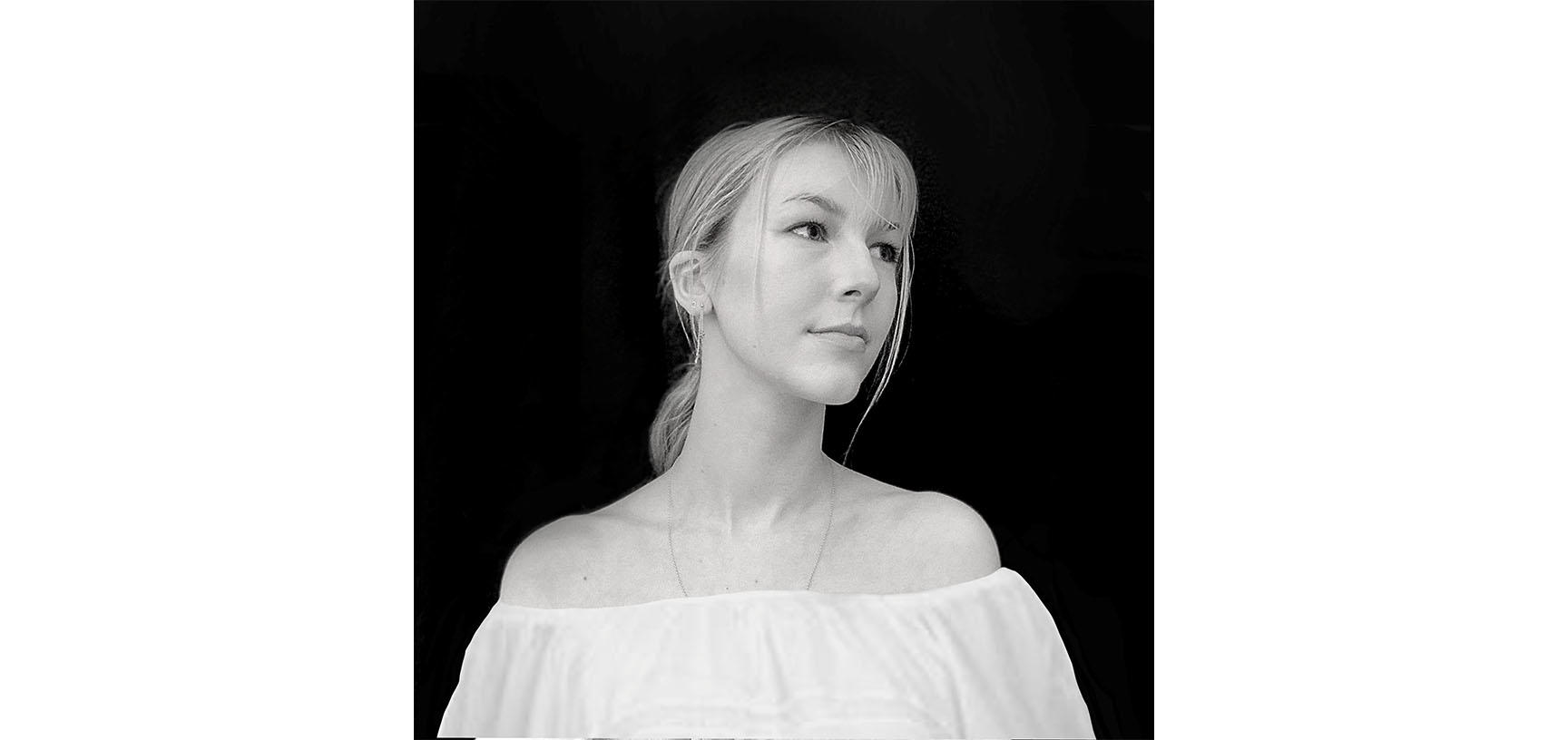 A woman wears a white top in front of a black background. She is looking over her left shoulder.