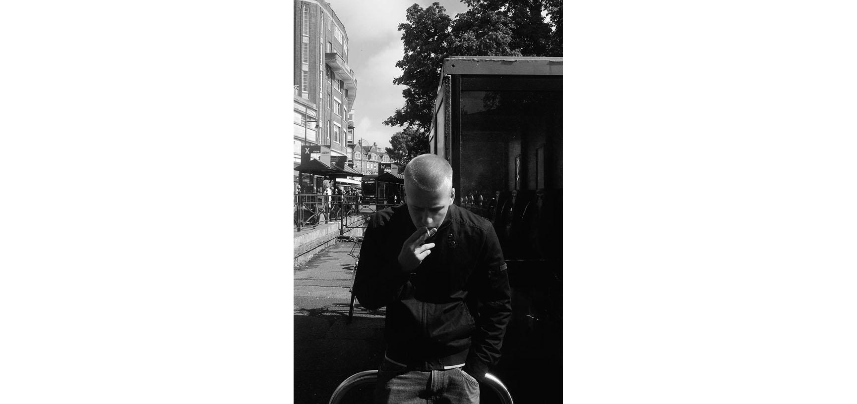 A black and white photograph of a man standing, head down, smoking a cigarette