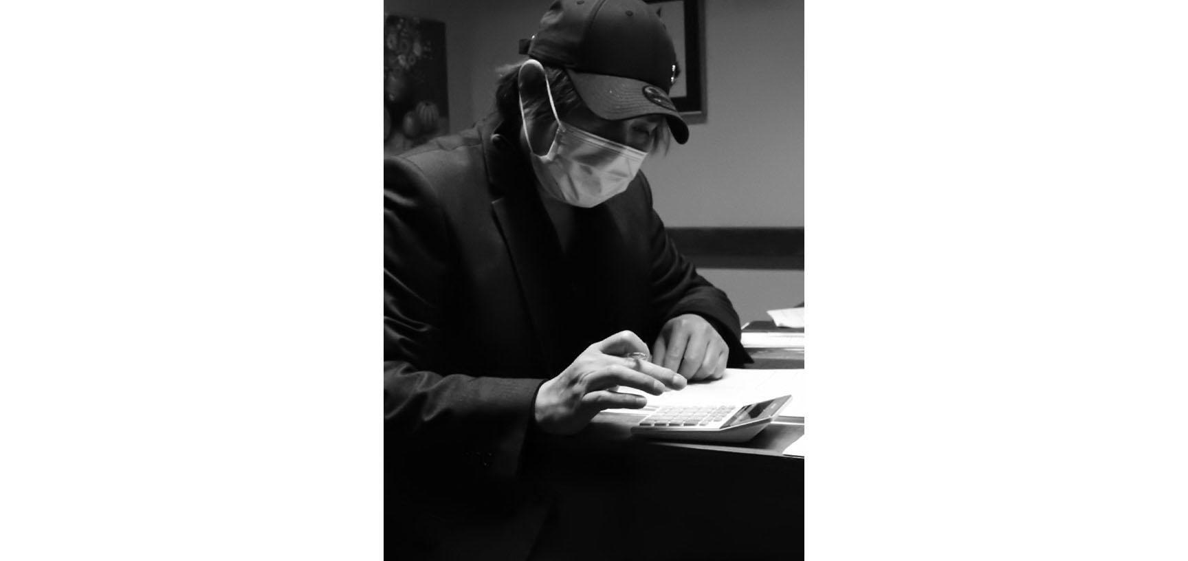 A black and white photograph of a man sitting at a table. He is about to write in a notebook. He wears a baseball cap on his head and a face mask across his mouth.