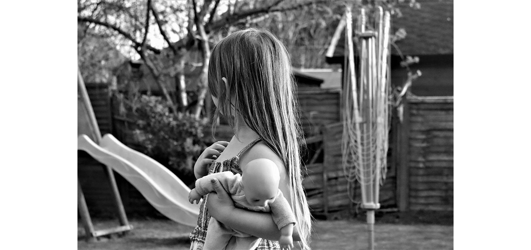 A black and white photograph of a young child. They are looking away from the camera towards a slide in the background. They hold a doll in their left hand.