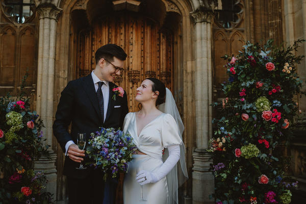 A couple stand in front of a wooden door with flower arrangements either side