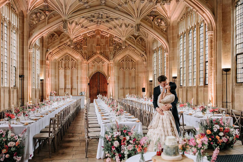 A couple kiss in front of three long empty tables in a large stone room